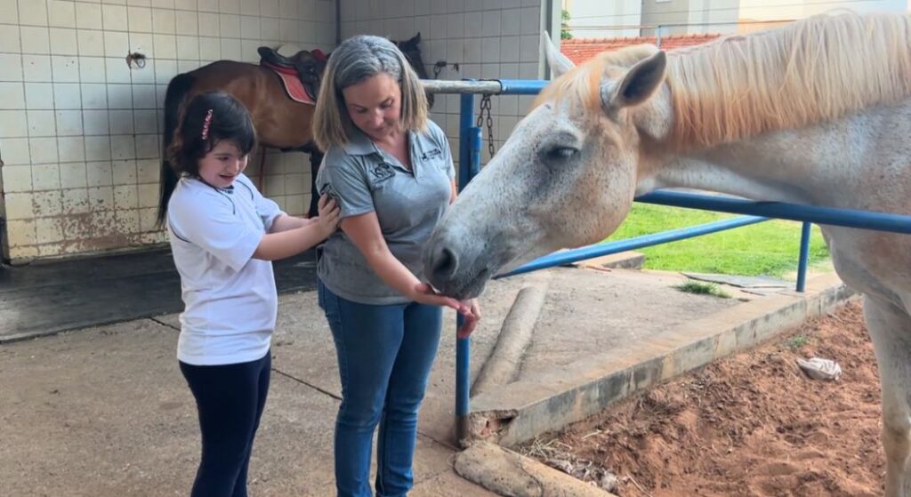 Tem uma menina observando uma cuidadora alimentar um cavalo