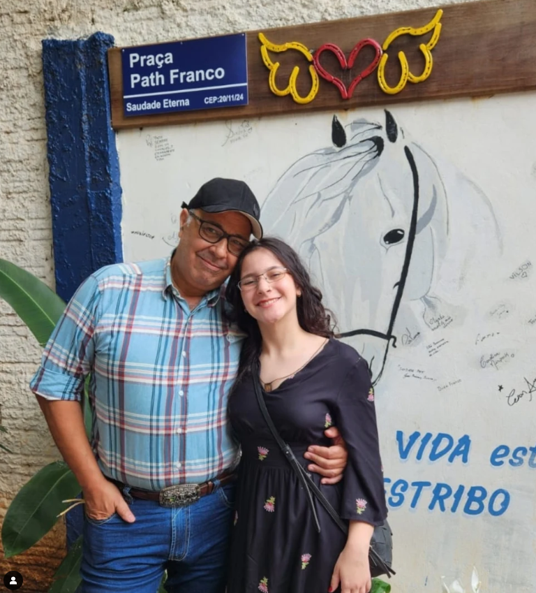 Um senhor e uma moça abraçados em frente a uma placa com o nome Praça Path Franco